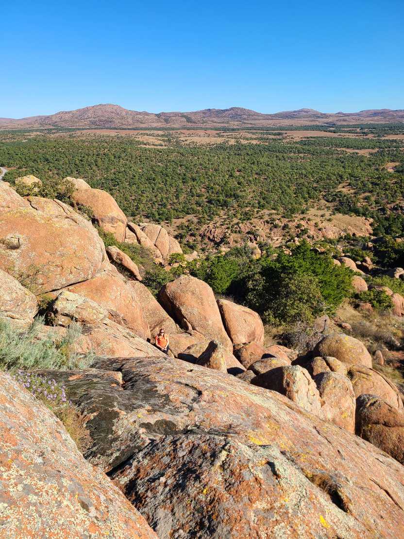 Large orange boulders with a view of greenery in the distance. Lydia sits on the rocks but is hard to see.