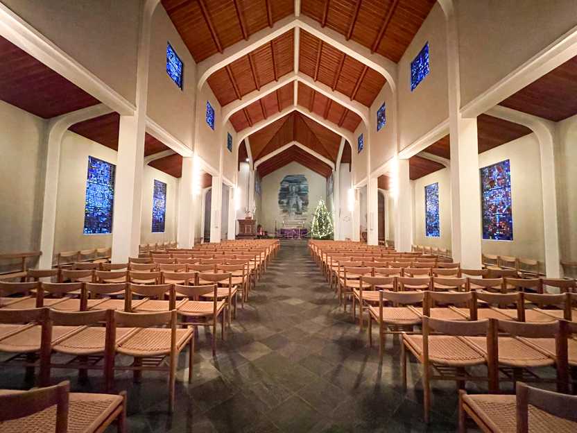 The interior of Skalholt Church.  The walls are white with a brown ceiling and brown chairs. There are stained glass windows on the walls.