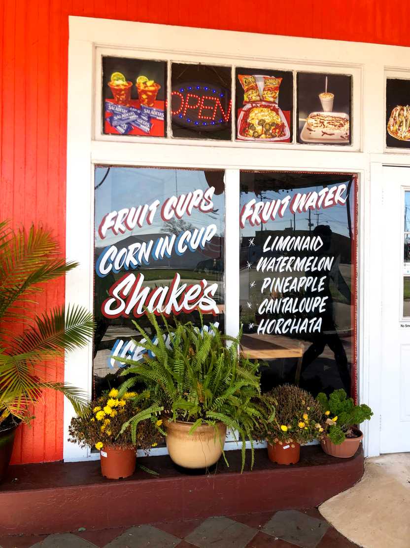 A red building with a white window frame. On the windows, there are lists of some of the offerings, such as "corn in cup," "shakes," "fruit water"