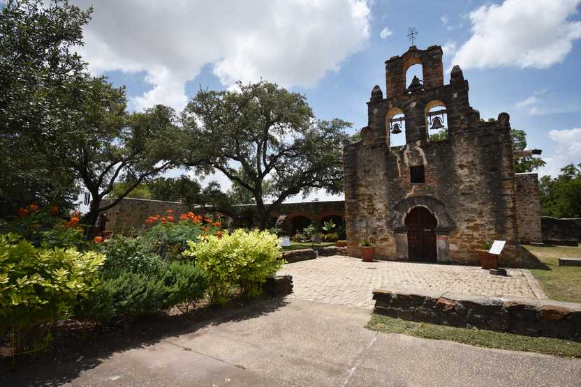 A small church with bells sits behind a beautiful garden of flowers.