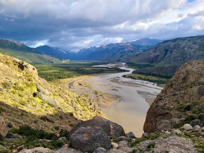 Looking down at a wide river next to a green valley. The river is framed by mountains in the distance.