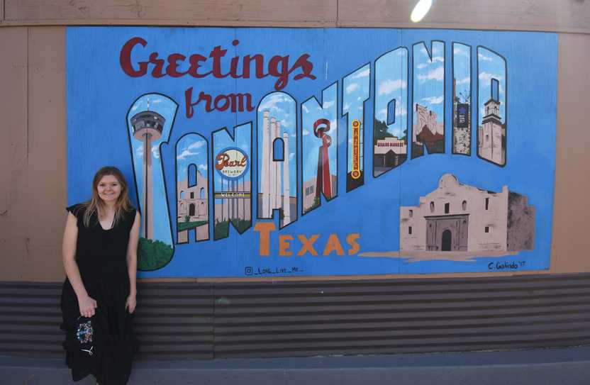 Lydia leans up against a mural that reads 'Greetings from San Antonio Texas'