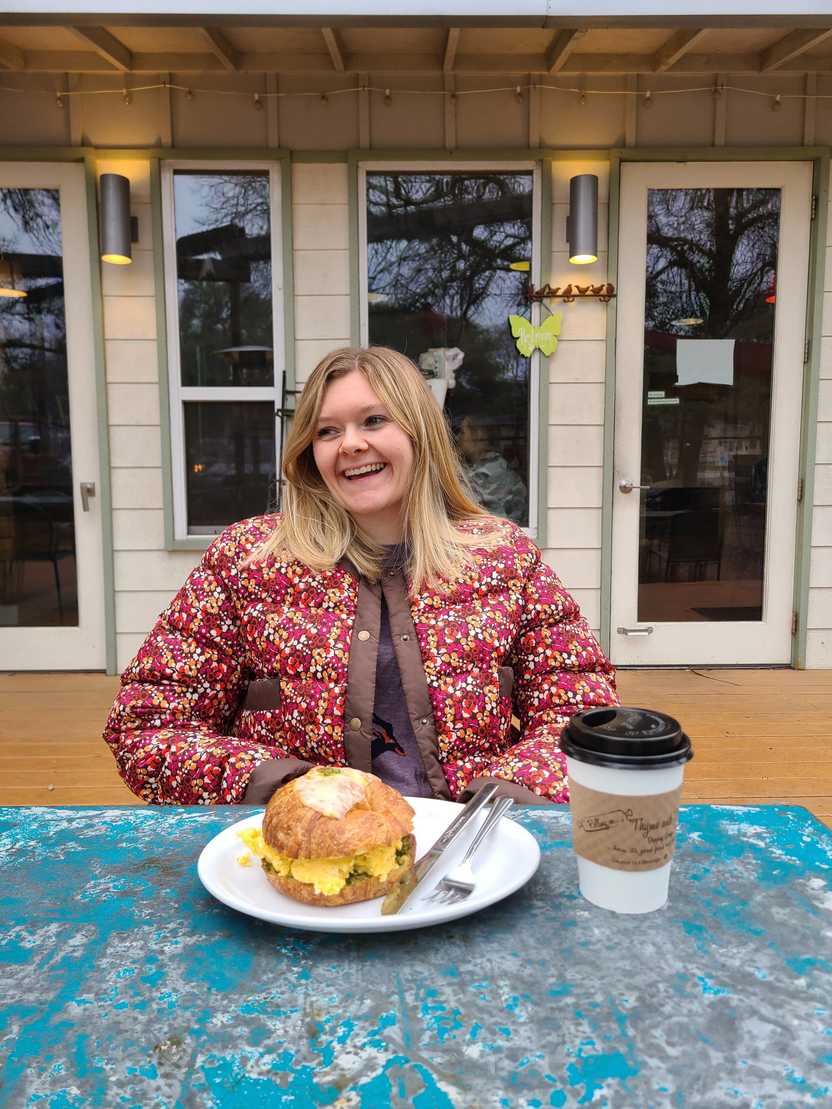 Lydia sitting at a table outside at Rolling in Thyme and Dough. On the table there is a a giant breakfast sandwich made with a croissant. You can see scrambled eggs between the bread.