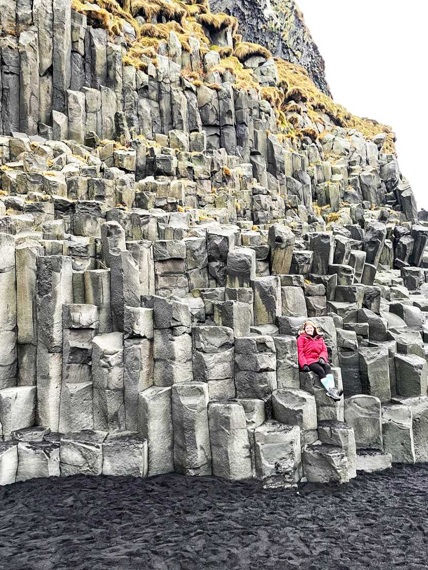 Lydia sitting on a wall of column-shaped rocks at Reynisfjara Beach.