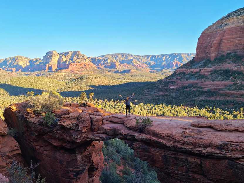 Lydia standing on Devil's Bridge and facing away from the camera. It is a giant natural bridge that made of orange rock. There are mountain views in the distance.