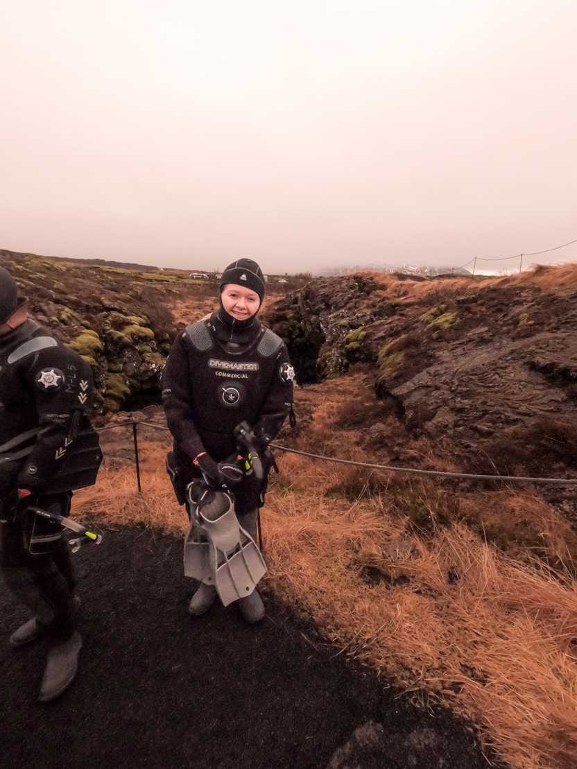 Lydia standing and wearing a dry suit holding a pair of flippers.
