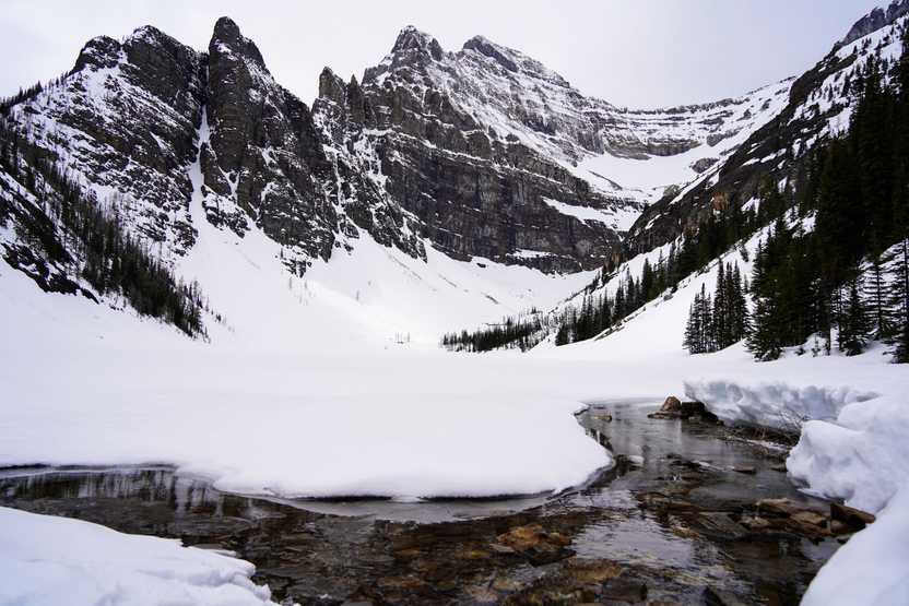 A partially frozen lake and mountains in the background. There is a couple feet of snow on top of the lake and mountains.