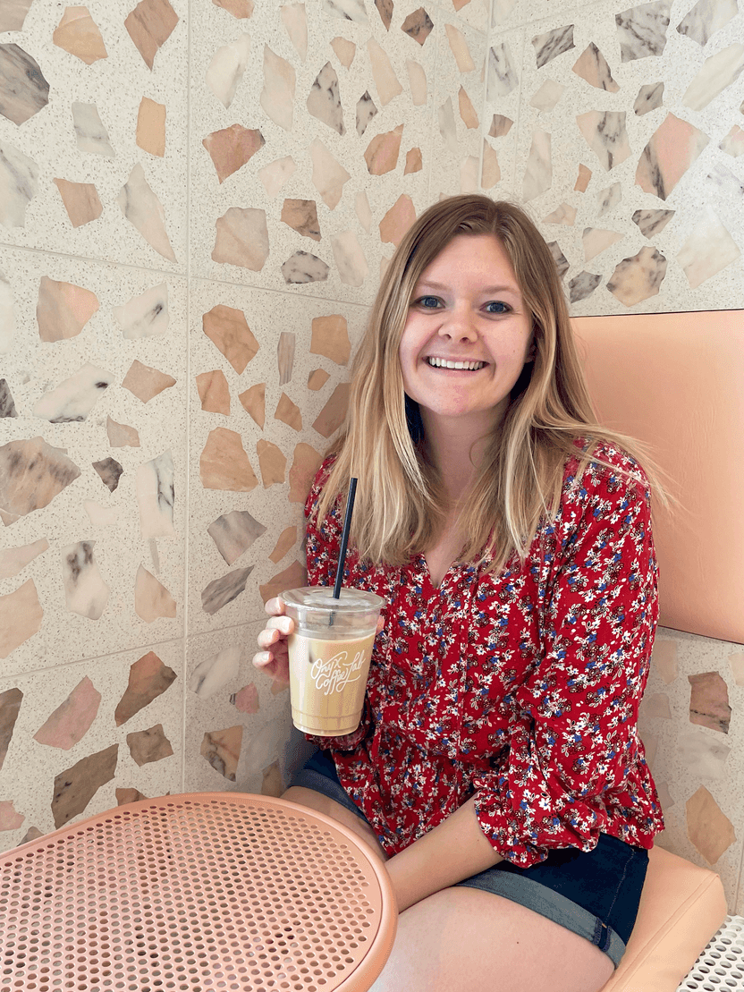 Lydia sitting at a pink table and holding a latte. The wall behind here has abstract geometric shapes.