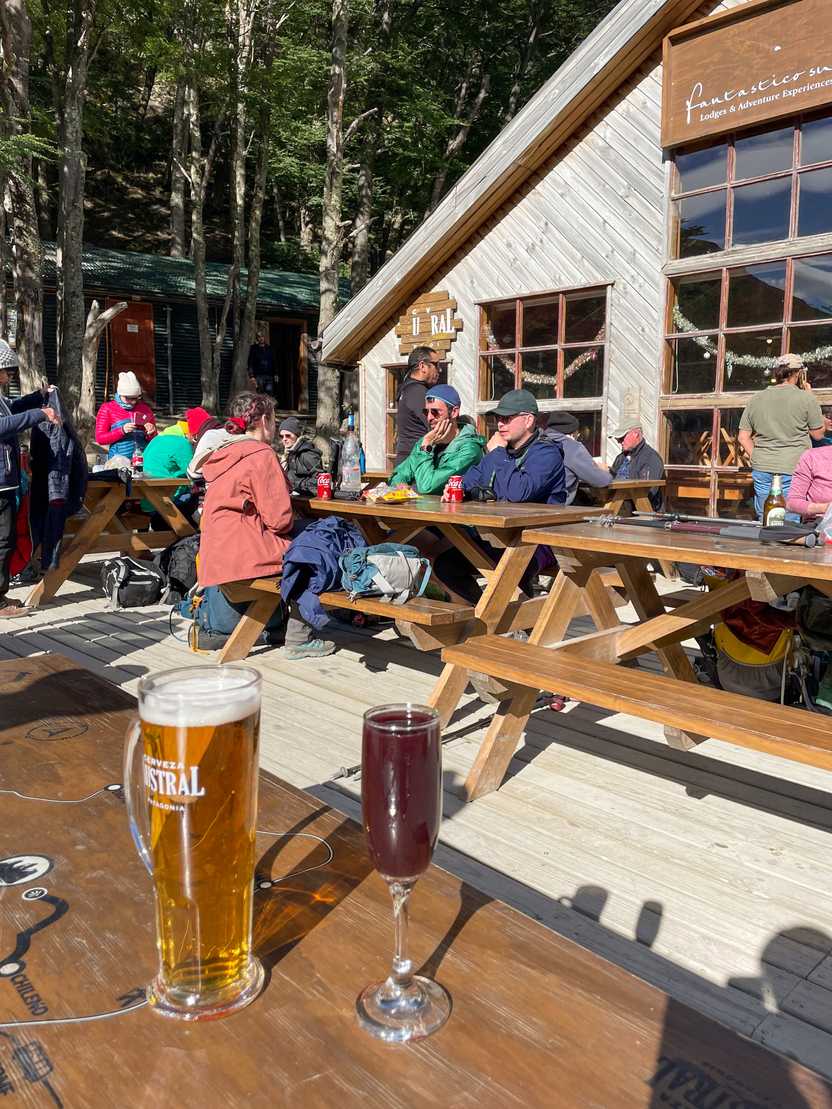 A beer and a glass with a purple drink outside on a picnic table at El Chileno. Several people sit at other tables in the background.