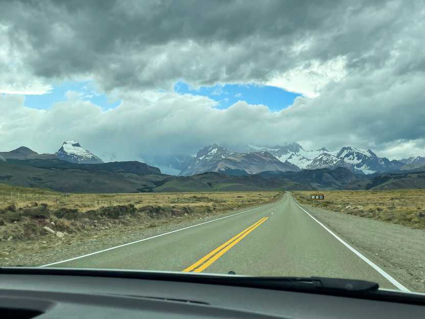 A long road that leads up an area of mountains partially covered in snow and ice. Clouds partially obscure the peaks of the mountains.