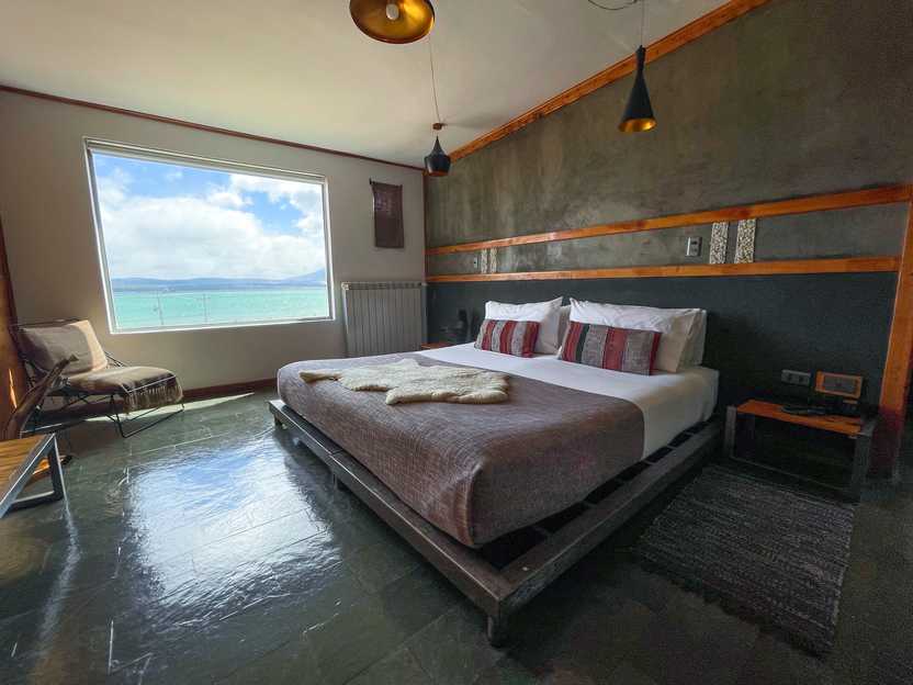 A hotel room with a view of an aqua colored lake. The bed has a brownish grey blanket and decorate pillows with red and grey stripes. There is a stripe of wood detailing across the middle of the wall and a chair in the corner.