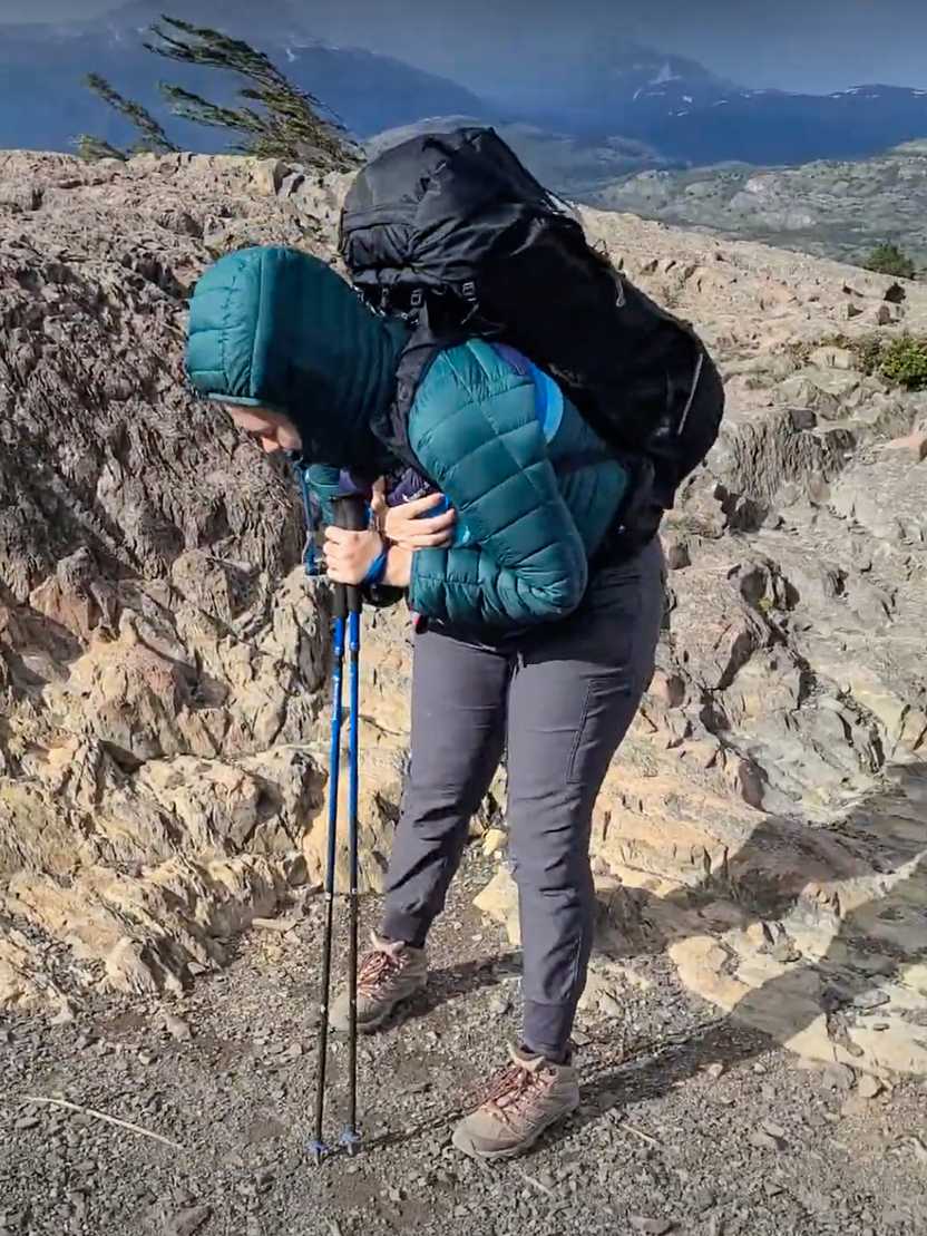 Lydia leading forward against the wind. She wears a green jacket and has her hood up. She is also holding trekking poles in one hand.