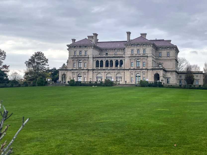 A large mansion across a green lawn. The building is three stories tall and has some arches over a balcony on the second floor. The building is tan and the roof is a dull reddish purple color.