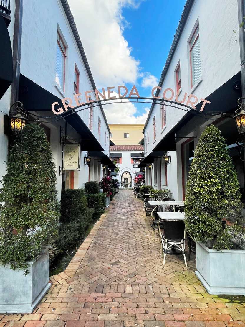 A alley way with white buildings on each side and the back. An arch above the alley reads "Greeneda Court." A few tables are on the side of the path.