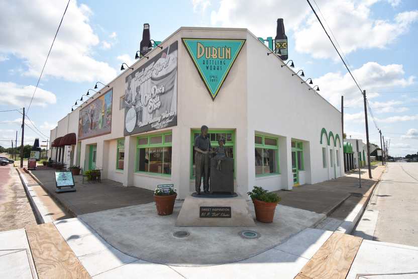 The exterior of Dublin Bottling Works front the corner. There is a statue in front of the building of W. P. Kloster, a triangle sign that reads "Dublin Bottling Works," a banner for Old Doc's Soda Shop and an empty sidewalk.