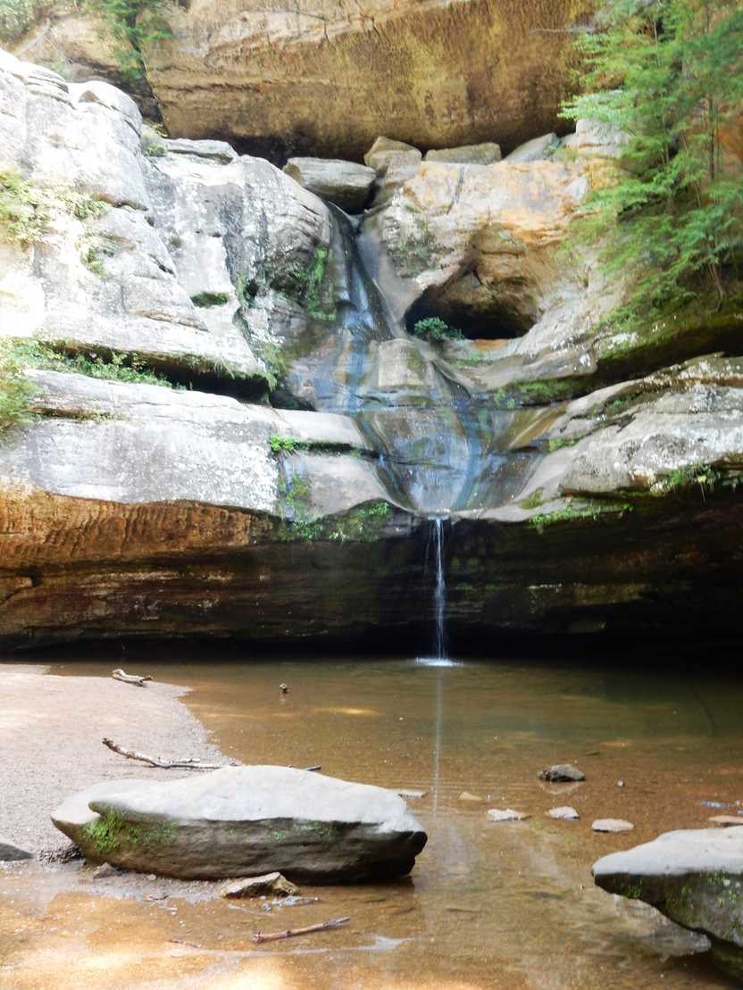 A view of Cedar Falls with a low water flow. Water trickles from a rock face into a pool of water.