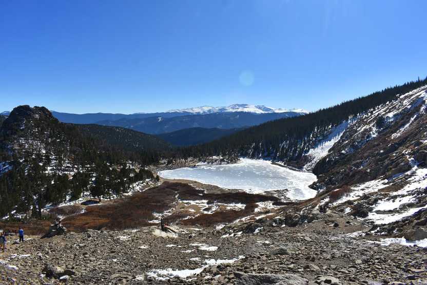 A view of the frozen St Mary's Lake from up on the hill above it. There is a little bit of snow on the ground and mountains in the background.