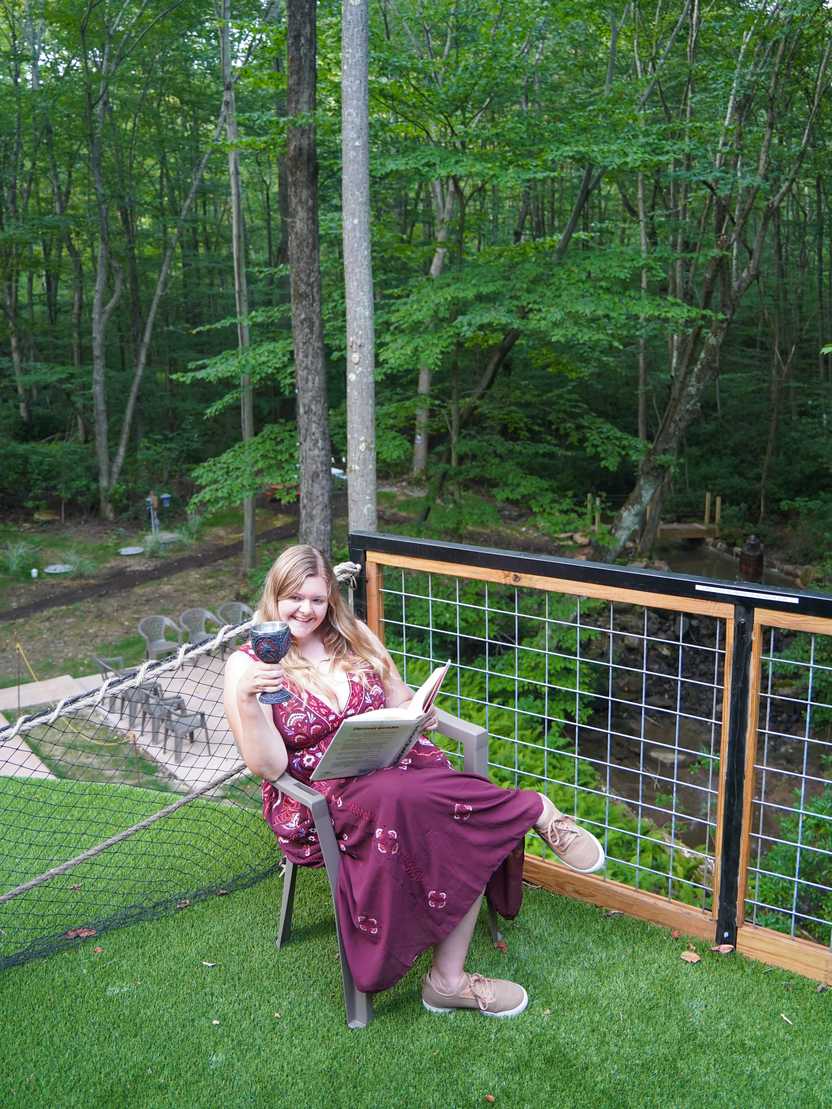 Lydia sitting on the balcony at the Hobbit House. She is reading a large book and holding a wine glass that resembles a goblet.