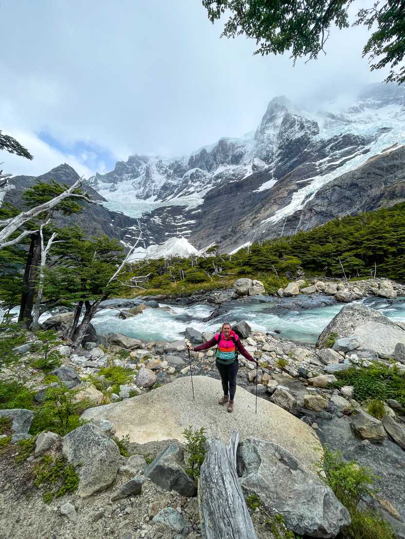Lydia standing on a rock with a view of the French Valley in the background. A river is stunning by and mountains clouded by fog are across the stream.