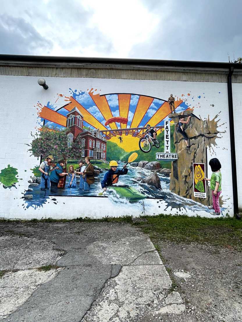 A colorufl mural that includes someone kayaking on a river, a biker, a rock climber, people playing music while standing in a river and someone parachuting from the New River Gorge Bridge.