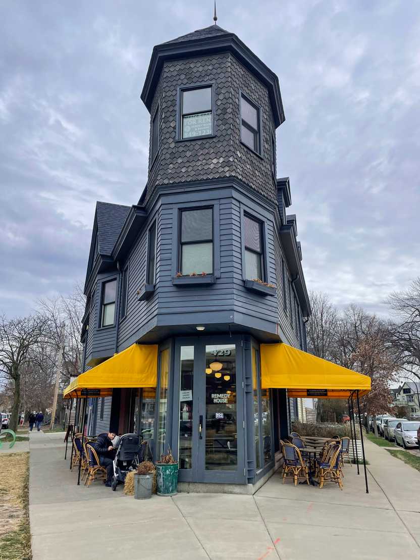 A narrow blue building on a corner with yellow awnings and outdoor tables on the sidewalk.