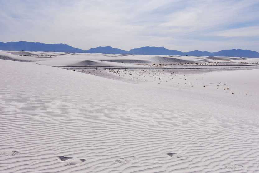 An expansive view of sand dunes with blue mountains in the distance. There are a few footsteps in the sand nearby.