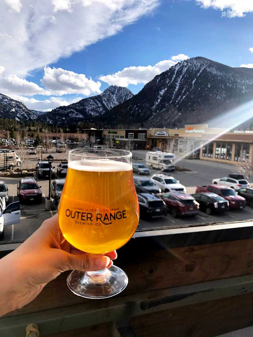 Holding up a glass of golden beer on the deck of Outer Range. There is a snow covered mountain in the distance. Holding up a glass of golden beer on the deck of Outer Range. There is a snow covered mountain in the distance.