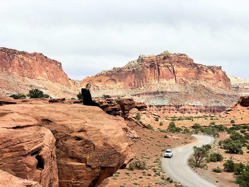 Lydia sitting on a rock face at the Panorama Point. There is a curving road below with a car driving.