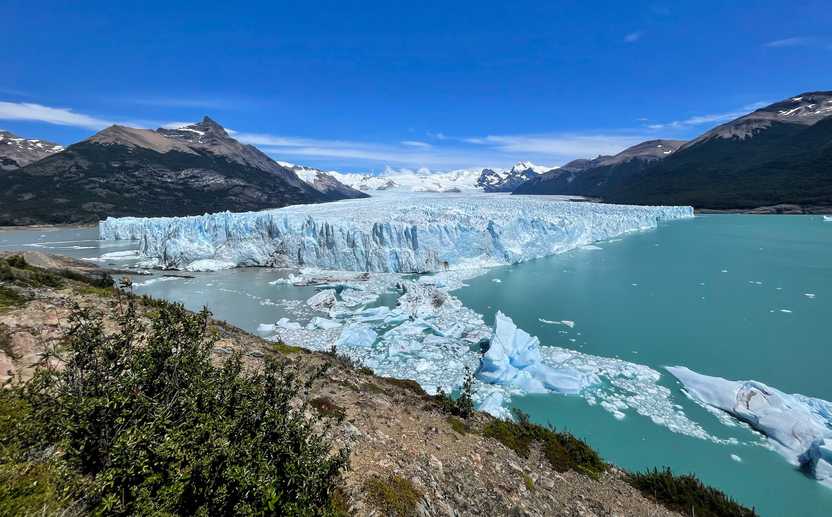 A wide view of a giant glacier on the water. There is some ice extending out from the center of the water. The water on the right side is a brighter green then the left side.