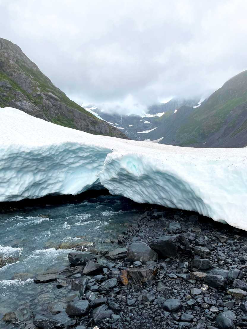 A small glacier with a river flowing under it and mountains in the background.