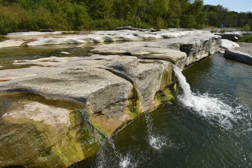 A close up view of the Lower Falls. Three small waterfalls come from the rocky cliff, which is about 8 feet tall above the water.