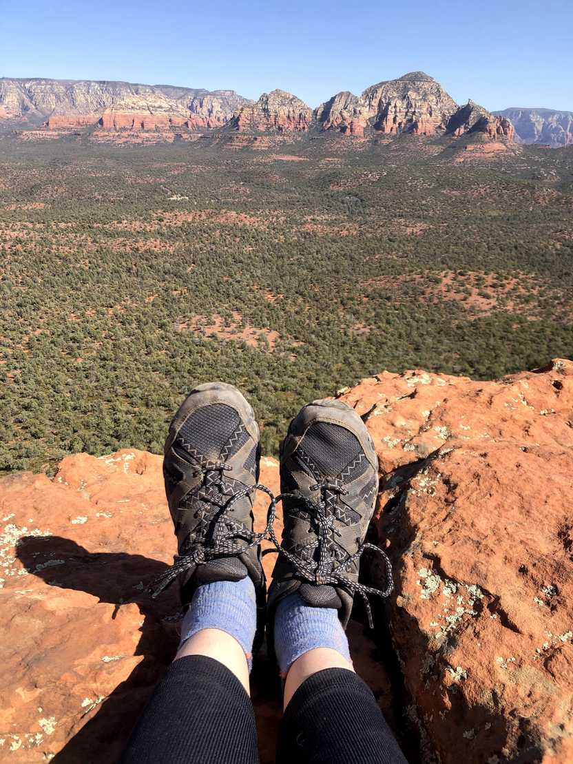 A photo of Lydia's feet wearing Merrell hiking shoes in Sedona. There are red rock mountains far out in the distance. A photo of Lydia's feet wearing Merrell hiking shoes in Sedona. There are red rock mountains far out in the distance.