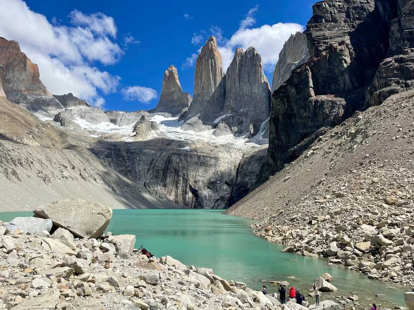 Looking across an aqua blue colored lake at the three towers inside of Torres Del Paine National Park. A few people are enjoying the view in the bottom right corner.