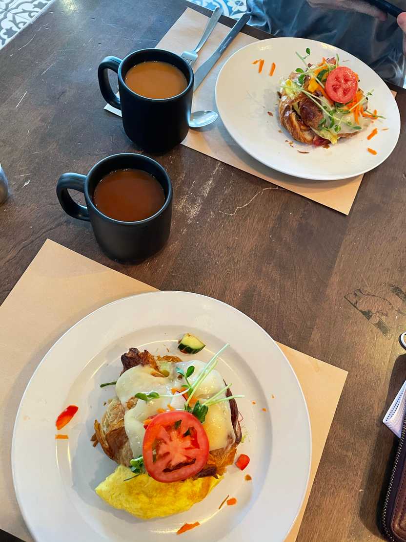 Croissant sandwiches topped with cheese and garnished with herbs and tomatoes. You can see some scrambled egg coming out of the sandwiches. There are also mugs full of coffee on the table.