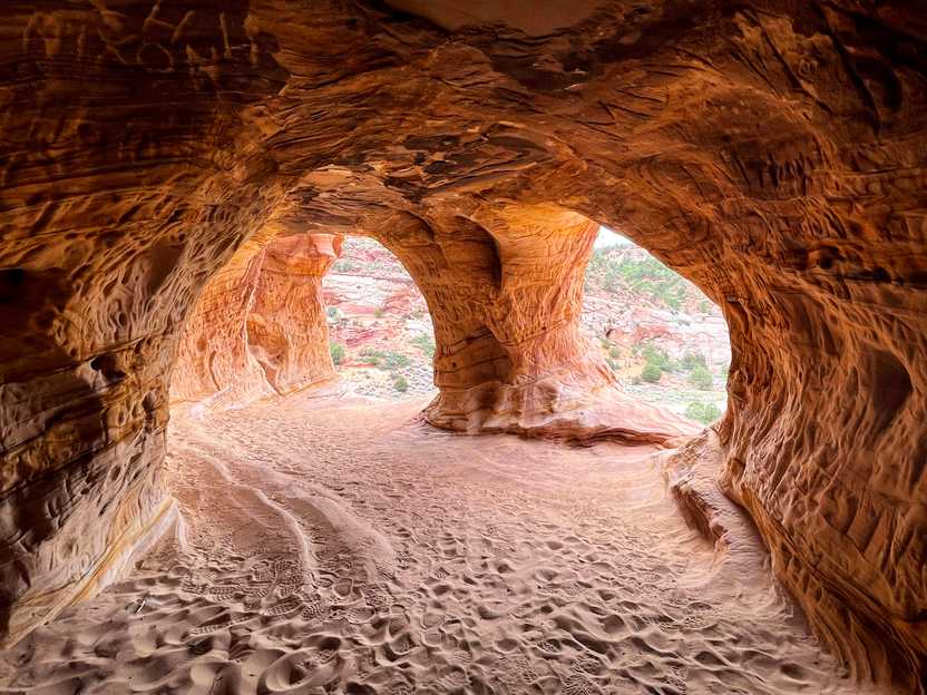 A cave area with sand at the floor and two openings. The walls of the cave are covered in carvings.