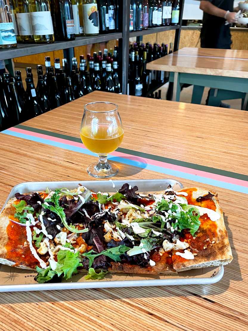 A flat bread pizza topped with lettuce and cheese. A glass of beer sits on the table next to it. The table is light wood with three stripes: one pink, one green, one blue.