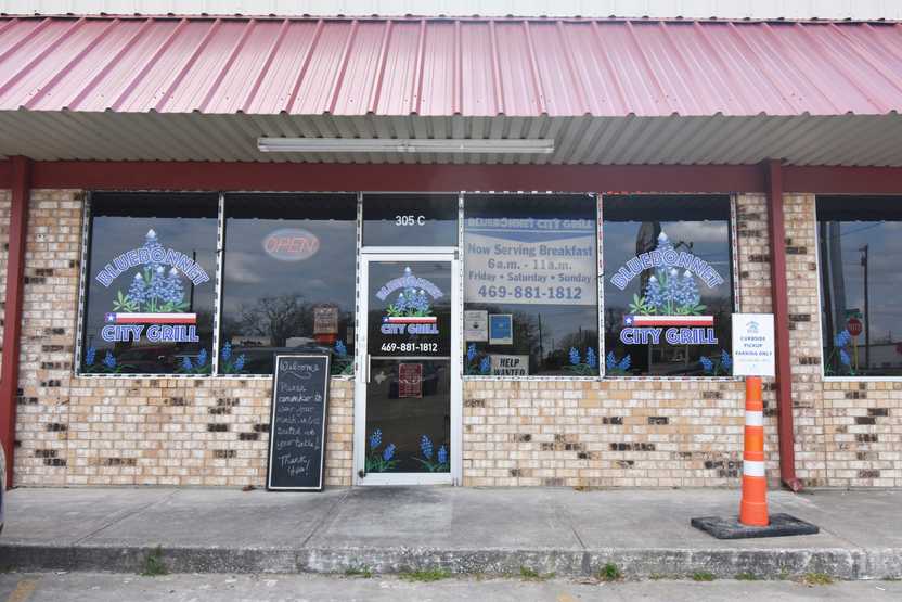 The exterior of Bluebonnet City Grill. There are bluebonnet flowers in the restaurant logo and a sign in the window about breakfast.