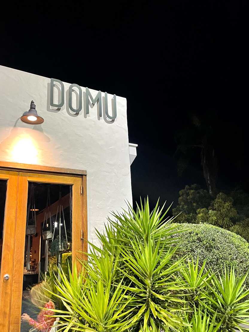 A white building with a sign reading DOMU with some green palms in front of it.
