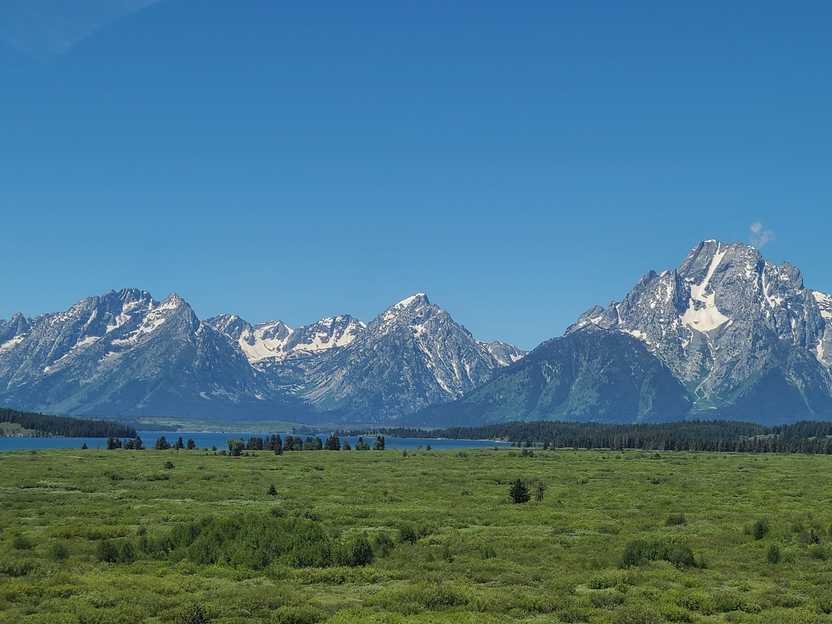 A view of mountains in the distance from across a valley. There is a valley of green grass and trees and a lake in the distance.