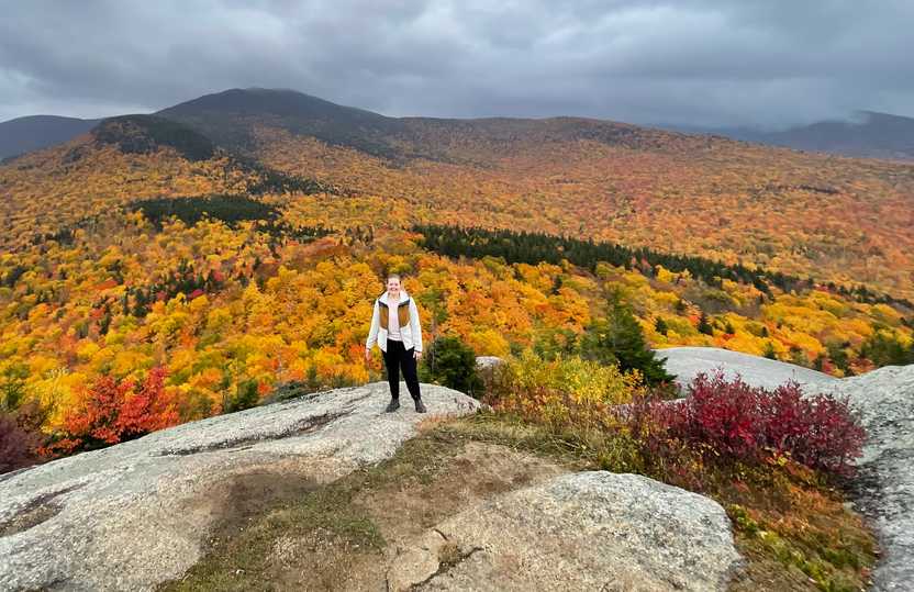 Lydia standing on a cliff with an expansive view of trees with bright foliage in the background.