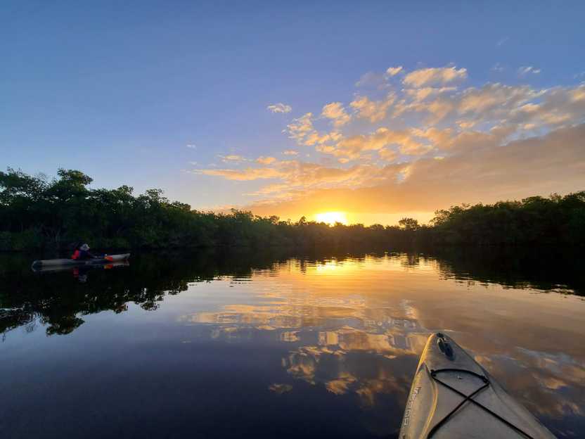 The front of a kayak with a sunrise in the sky and reflecting on the water. Yellow and orange clouds reflect onto the water