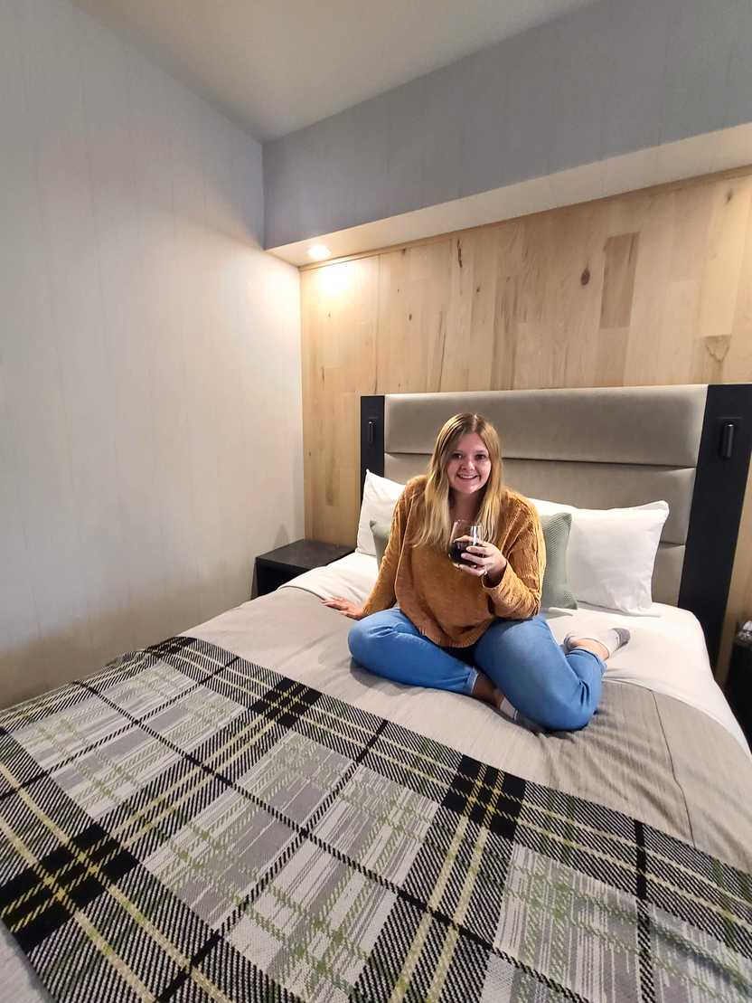 Lydia sitting on a bed inside the Peaks Hotel. There is a gray, plaid blanket on the bed.