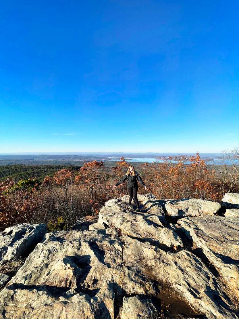 Lydia standing on a rocky area with view of trees and a lake in the distance in the background.