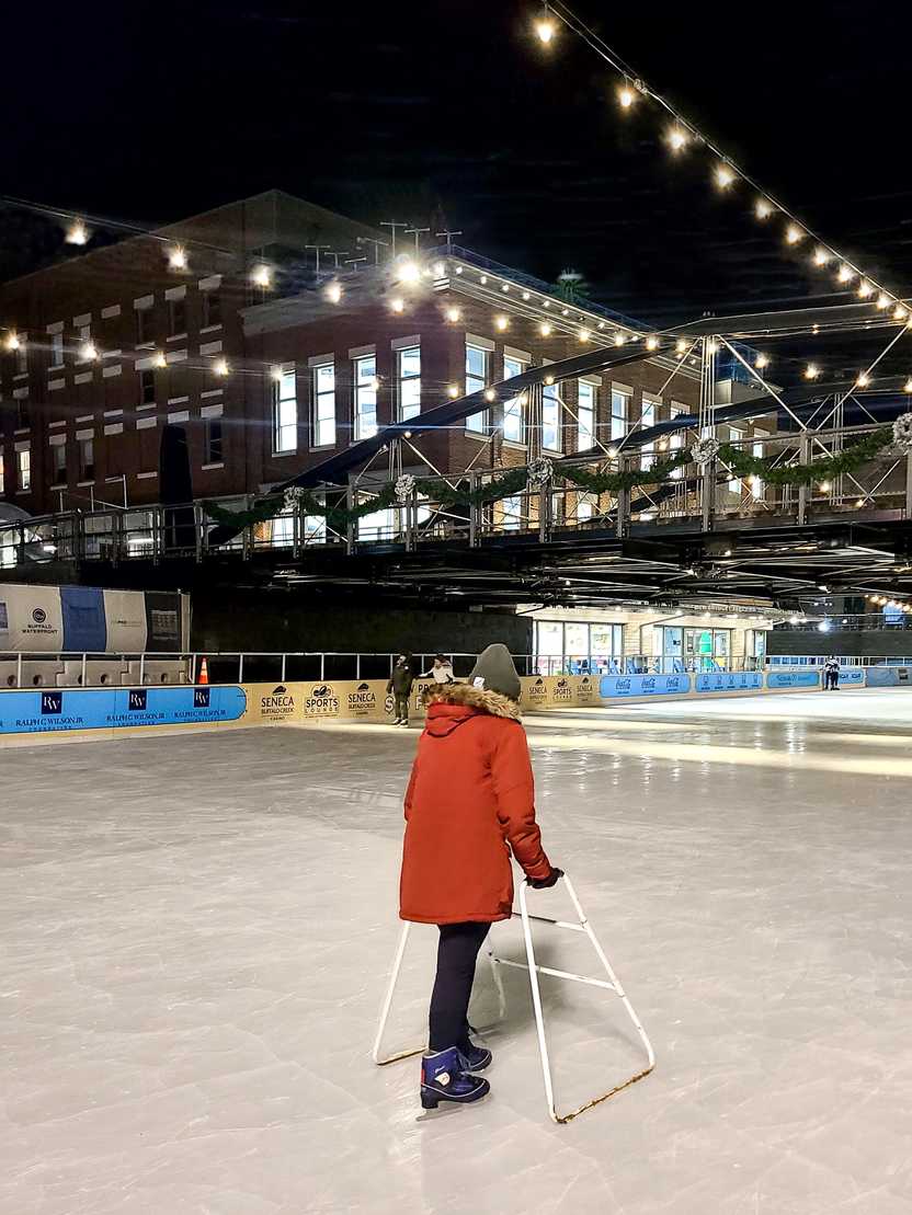 Lydia ice skating using a walker. There are hanging lights and a bridge above the rink. Lydia wears a red coat and green hat.