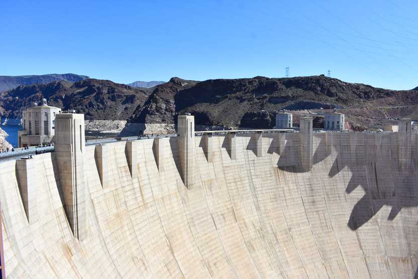 A view of the Hoover Dam. The dam is white and towers many feet. There are people walking across it.