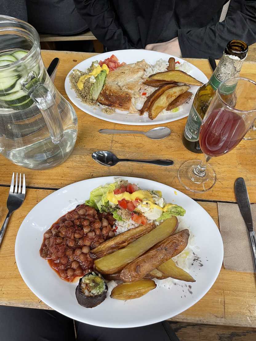 A plate of food that includes potato wedges, salad, baked beans and a sushi roll. There is a Calafate Sour cocktail next to the meal.