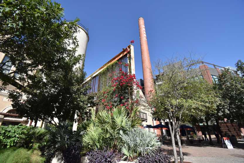 Industrial structures surrounded with trees and flowers. There is a tall smokestack.