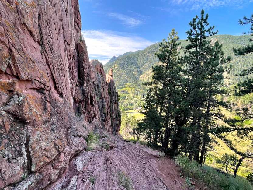 A red rock face with a trail next to it and green hills in the distance.