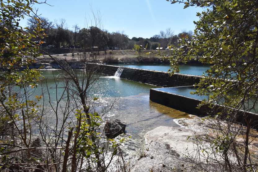 Blue water flows over a dam to create two levels of water. A stone wall holds in a pool of water on the right side of the view.