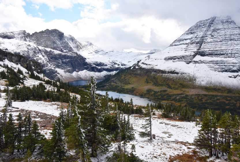 A blue lake with a mountain in the background. The ground surrounding the lake has snow and there are pine trees.